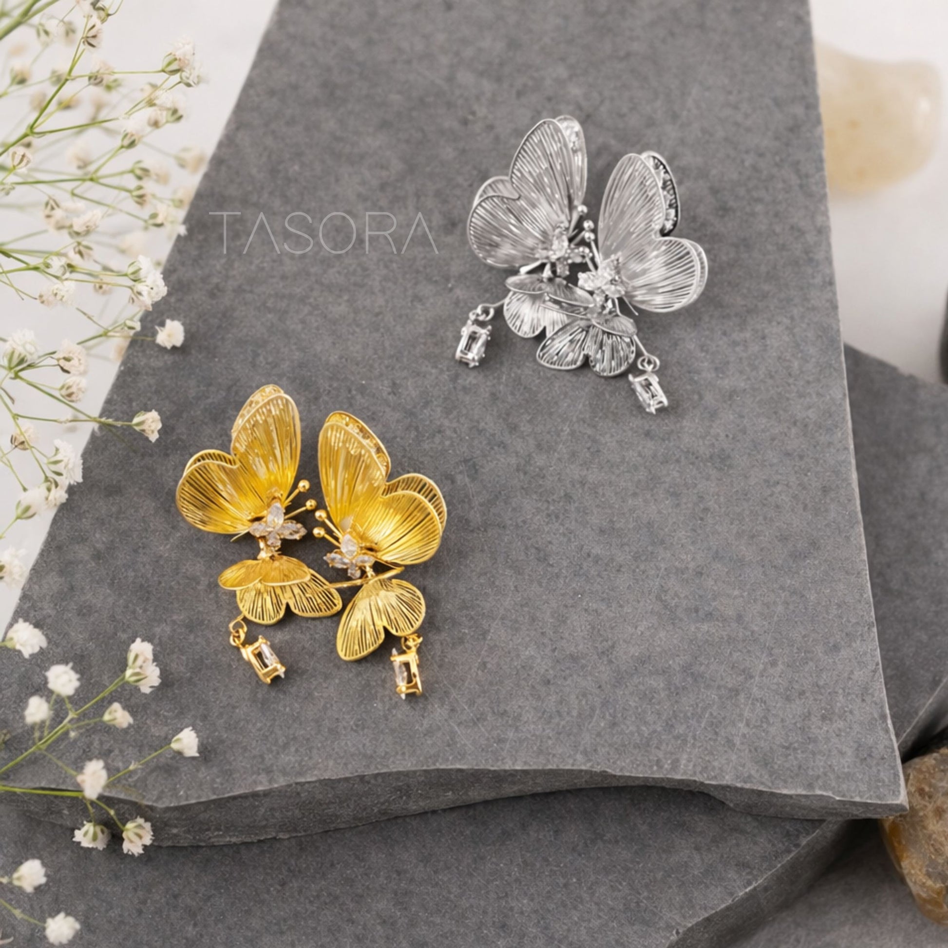 Butterfly-shaped earrings in gold and silver on a gray surface with 'TASORA' branding.