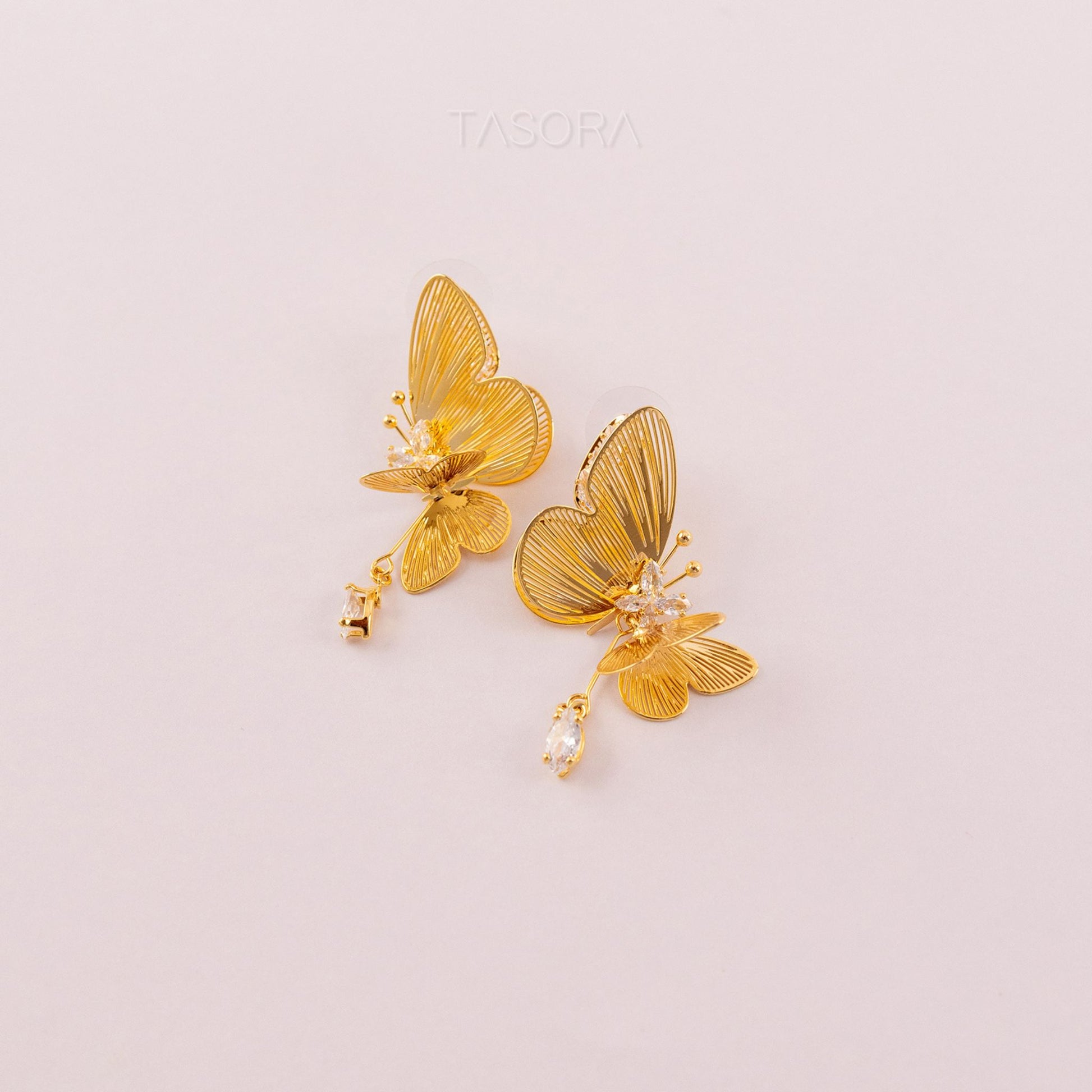 Golden butterfly shape Blinky Butterfly earrings on a light pink background with 'TASORA' branding.