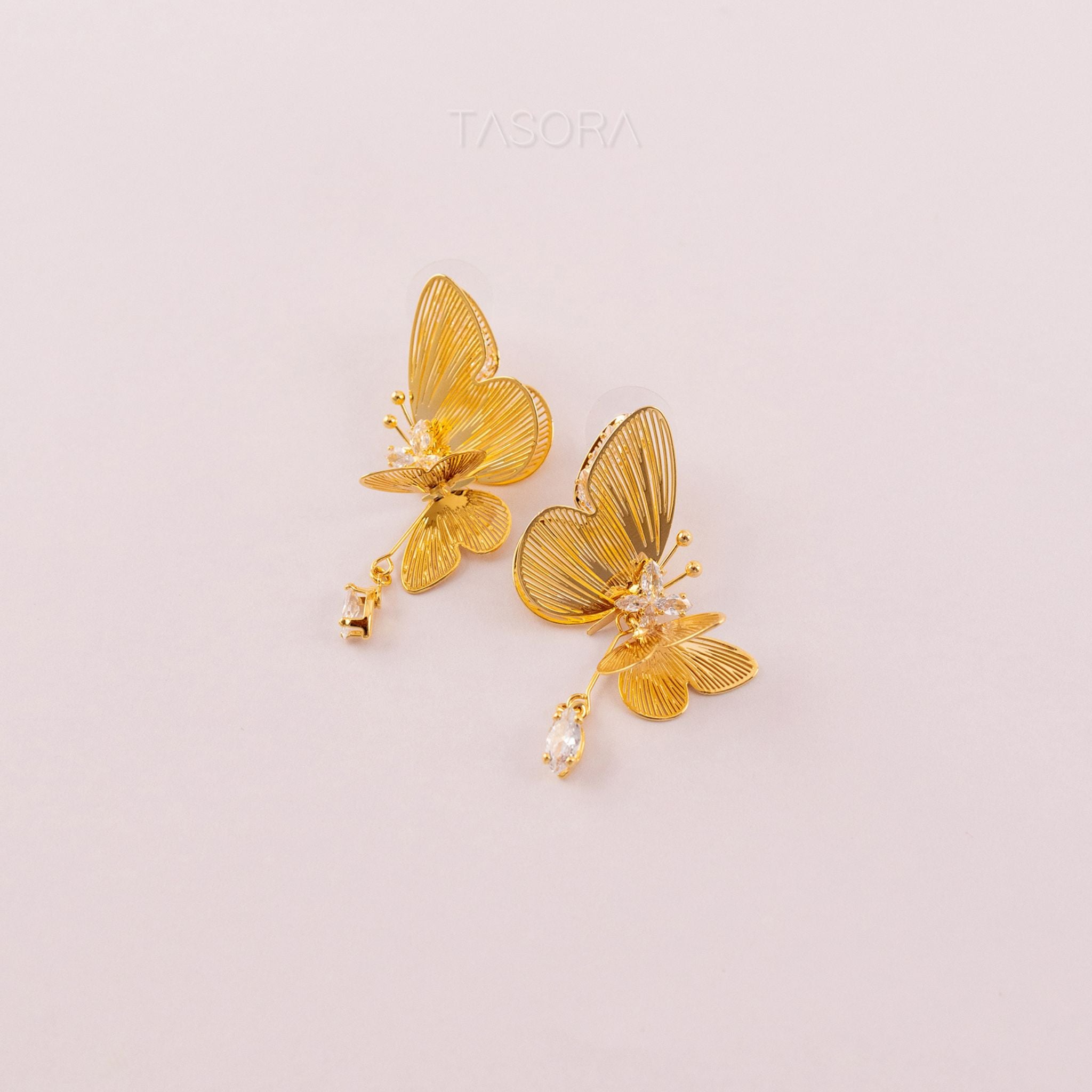 Golden butterfly shape Blinky Butterfly earrings on a light pink background with 'TASORA' branding.