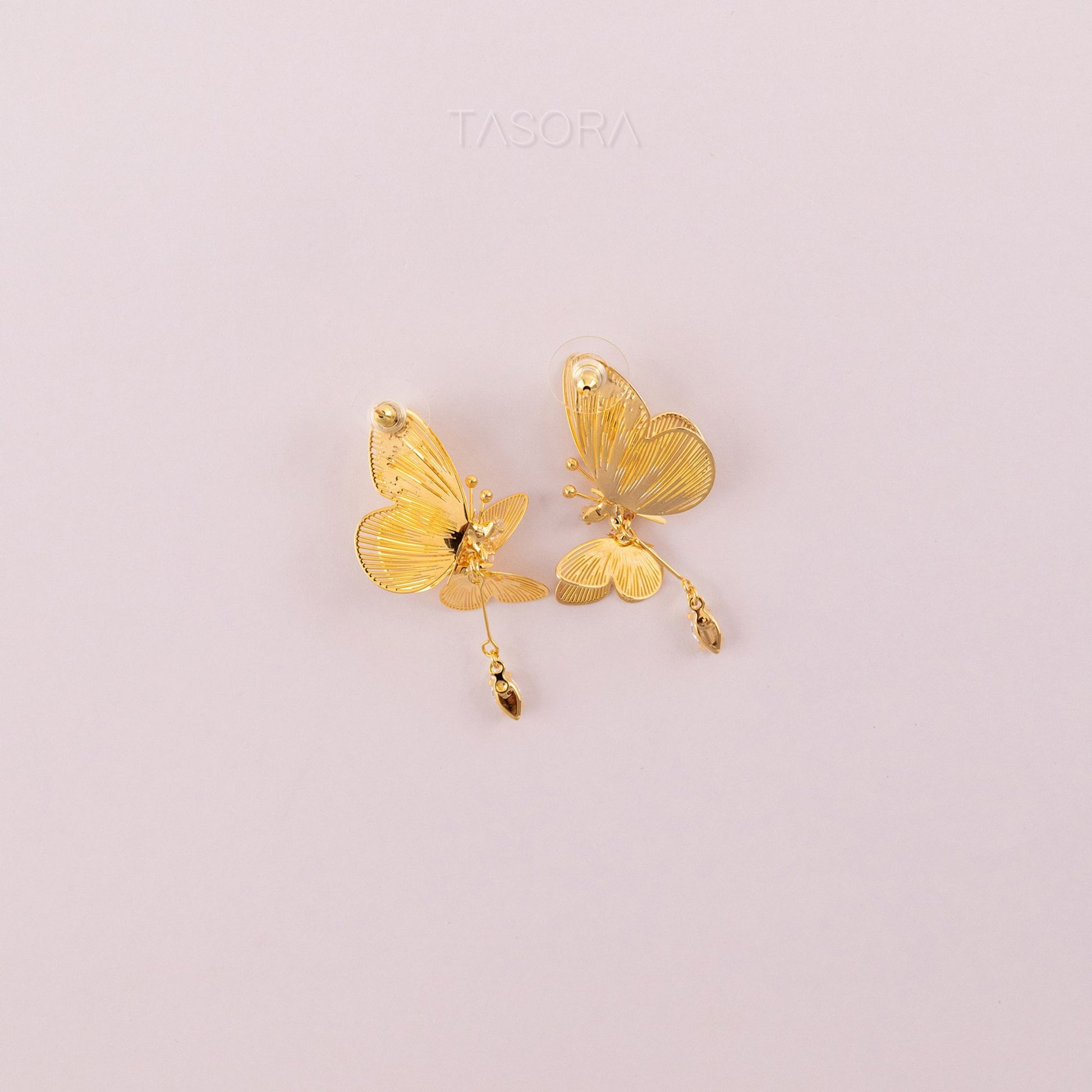 Back side of Golden butterfly shape Blinky Butterfly earrings on a light pink background with 'TASORA' branding.