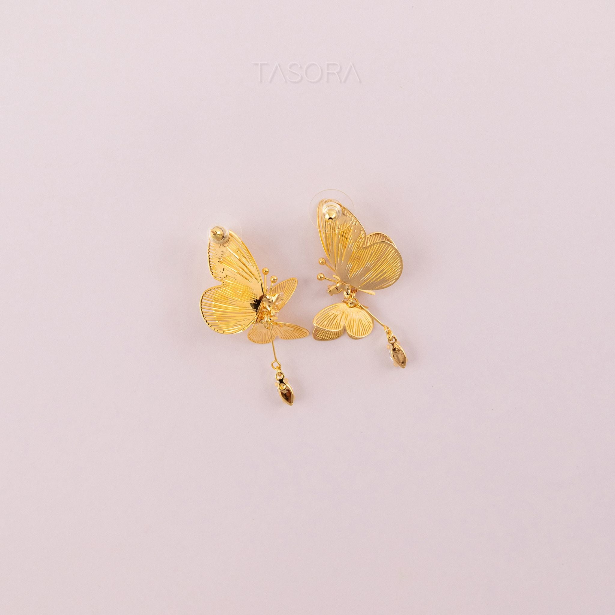 Back side of Golden butterfly shape Blinky Butterfly earrings on a light pink background with 'TASORA' branding.