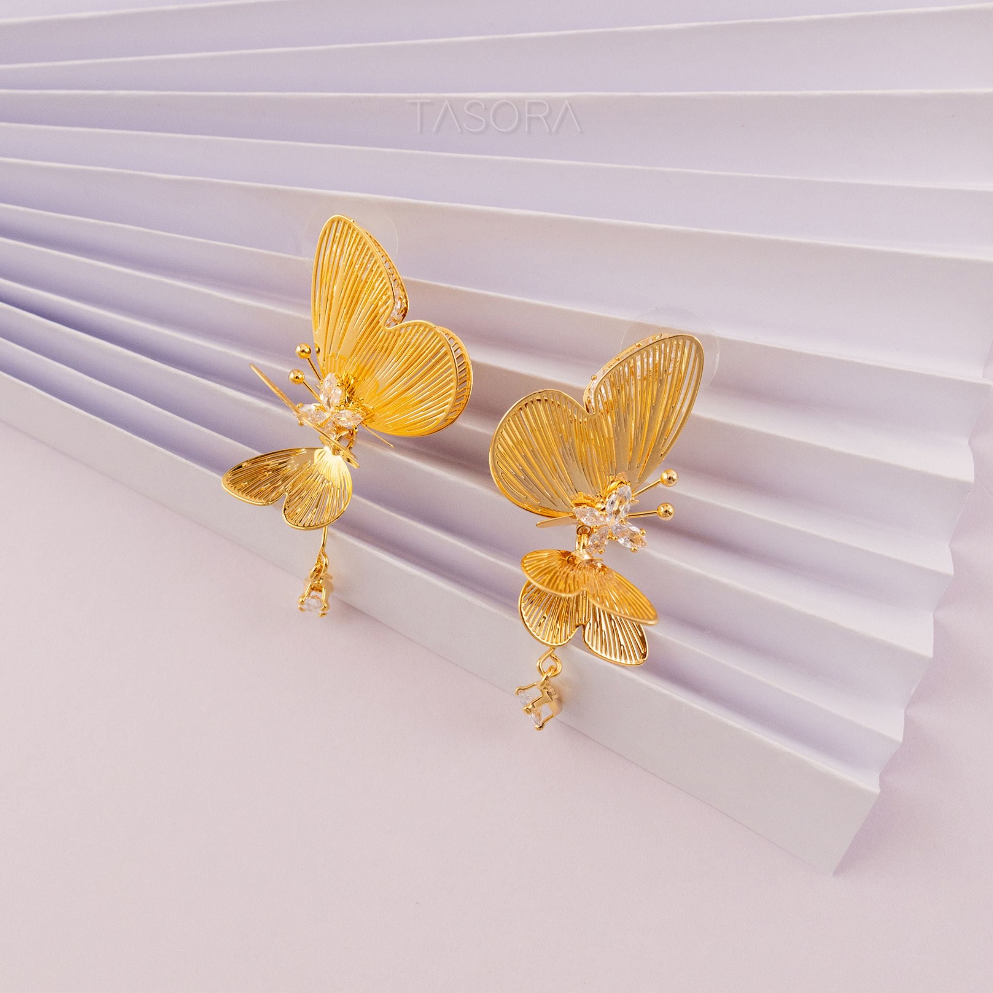 Golden butterfly shape Blinky Butterfly earrings on a white and purple striped background with TASORA branding