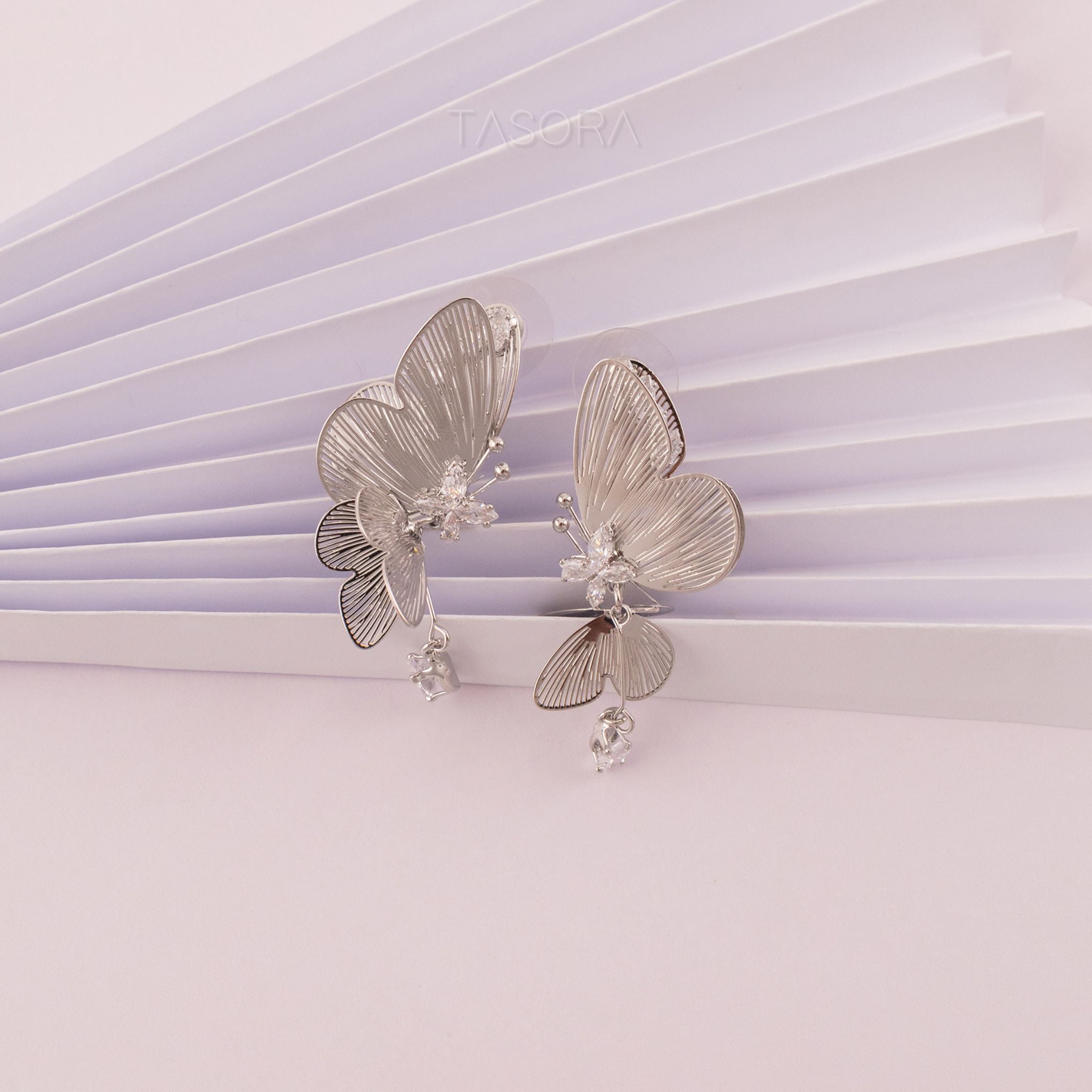 Butterfly-shaped Blinky Butterfly Silver earrings on a light purple texture background TASORA branding