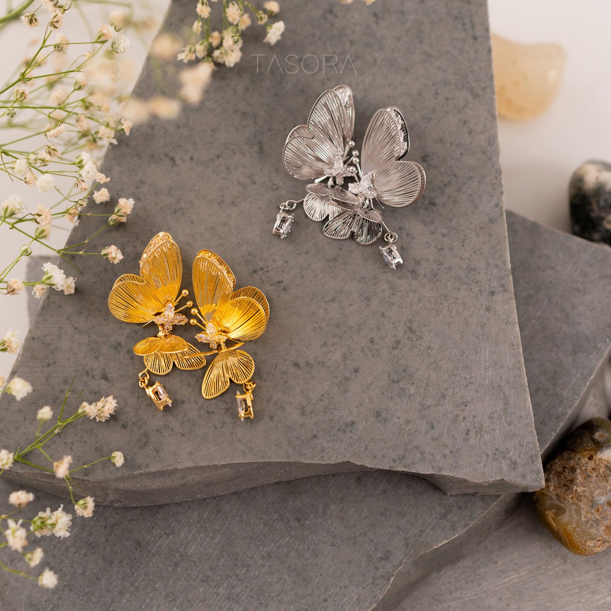 Butterfly-shaped Blinky Butterfly earrings on a stone surface with small flowers with TASORA brand visible