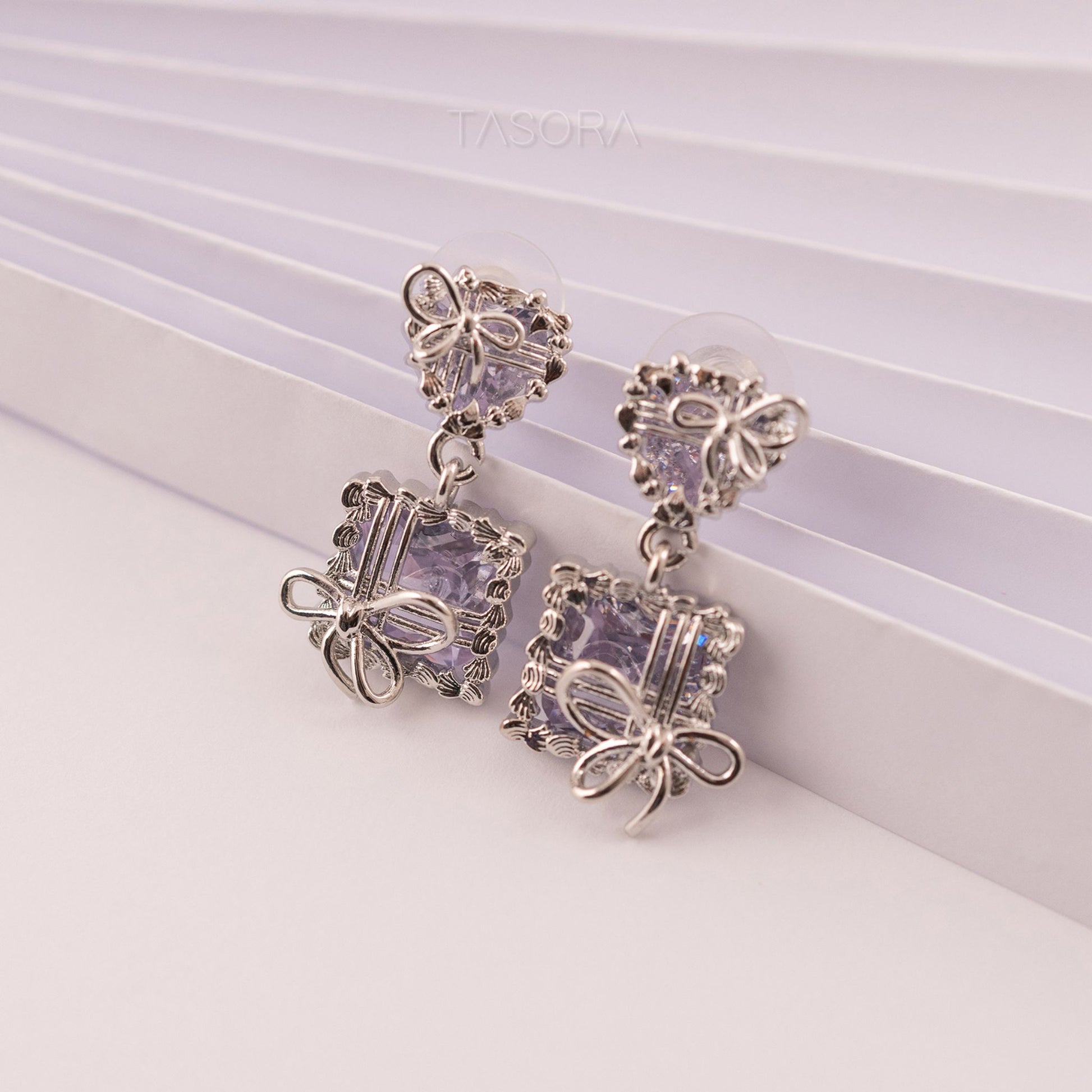 Pair of Bow Belle silver earrings with purple gemstones on a light background with TASORA branding