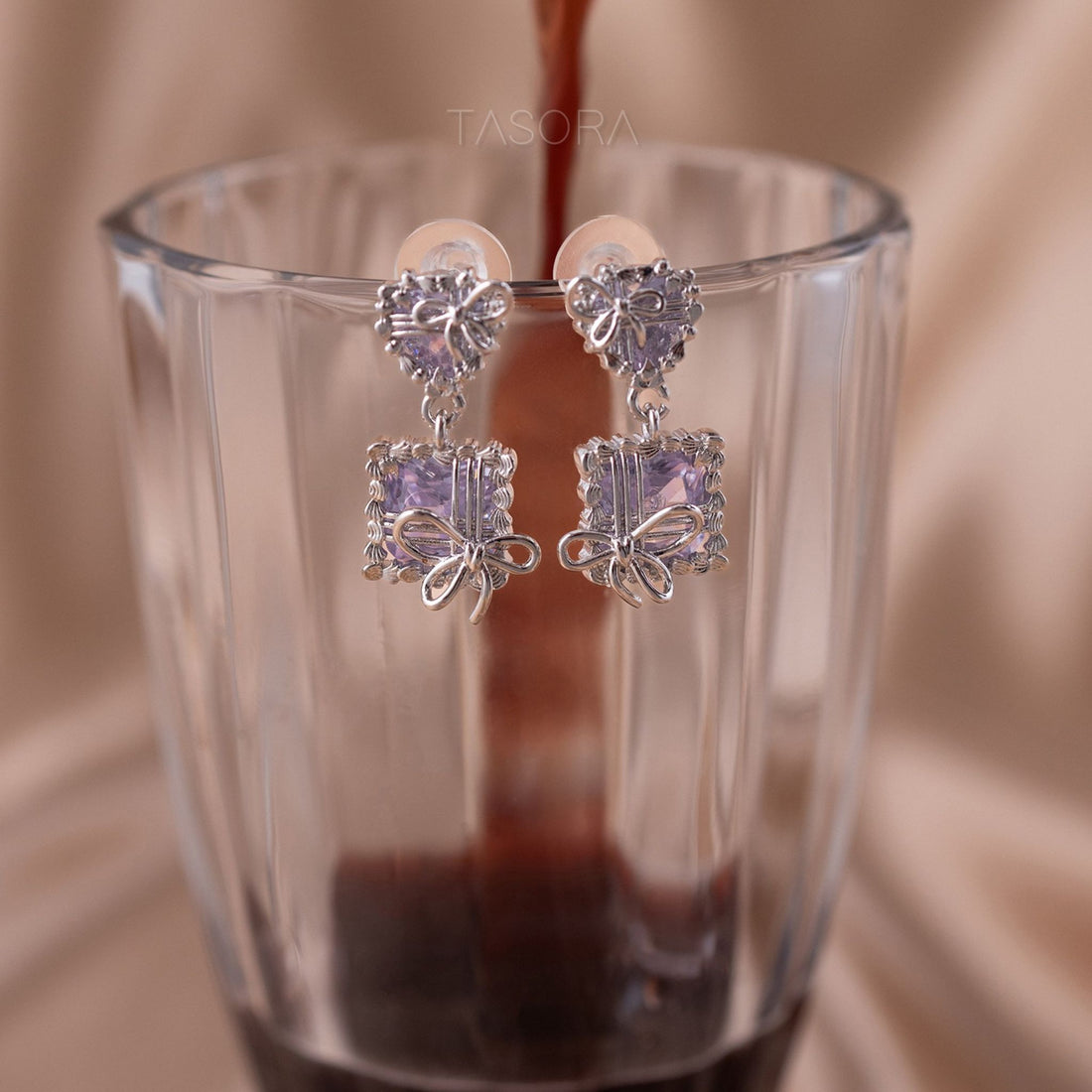 Pair of Bow Belle earrings by TASORA with purple gemstones in a glass of coffee.