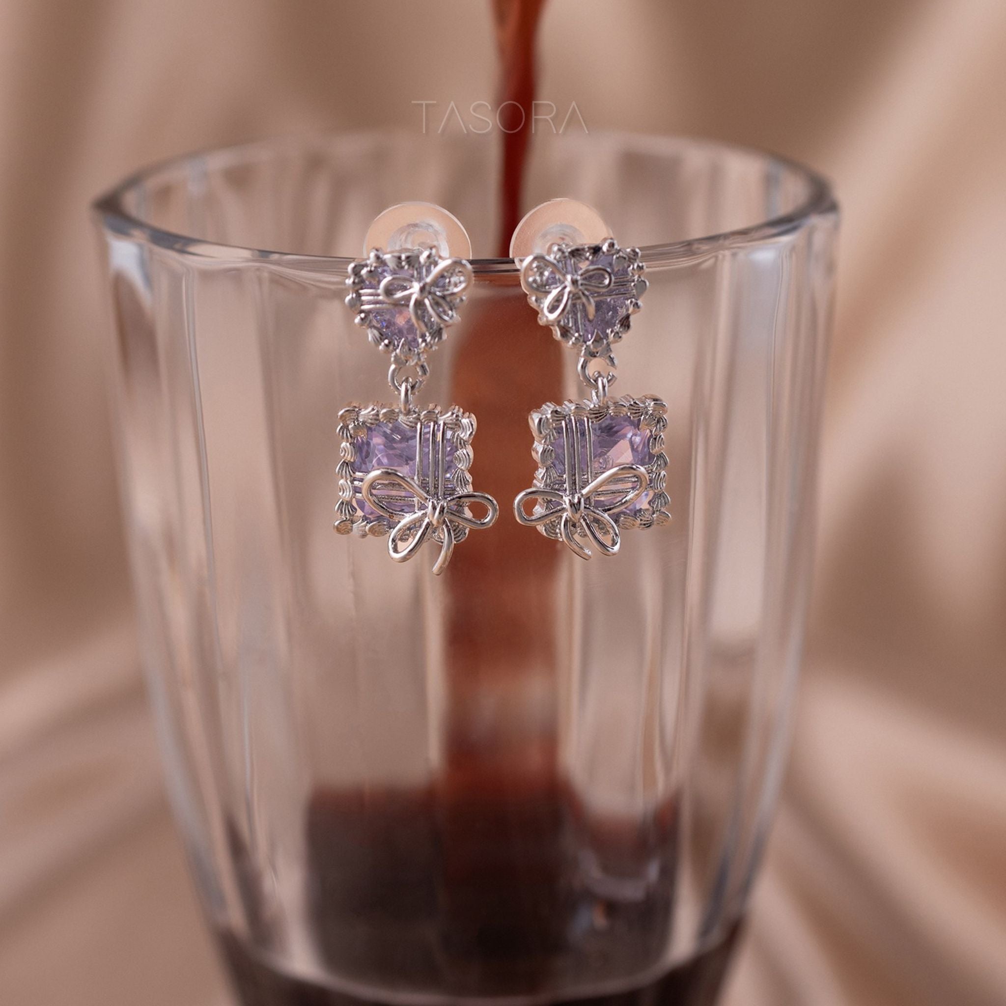 Pair of Bow Belle earrings by TASORA with purple gemstones in a glass of coffee.