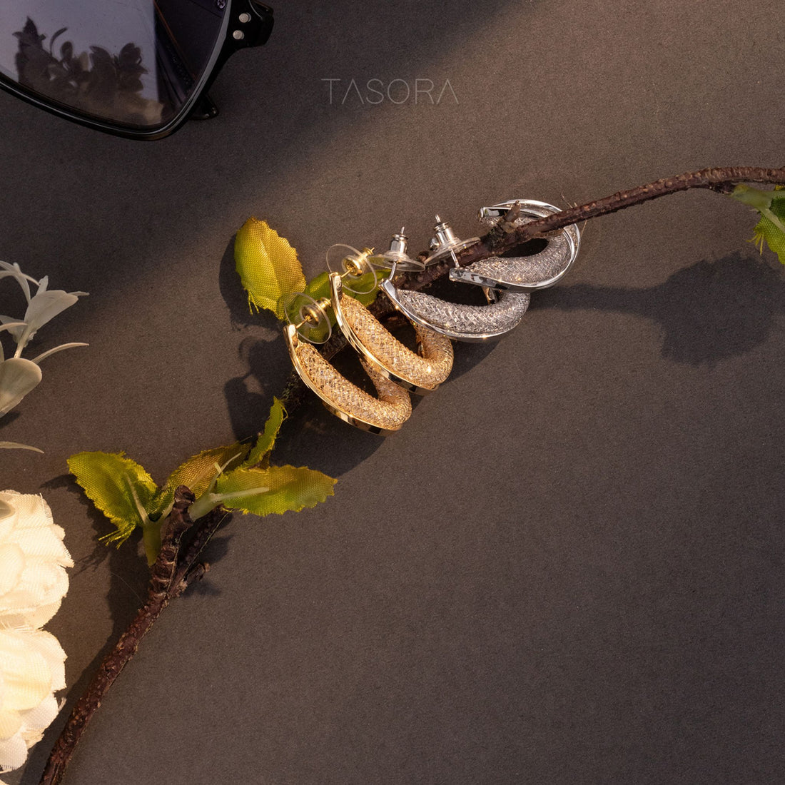 Pair of Curvy Hoops earrings on a branch with leaves against a dark background, featuring the brand 'TASORA'.