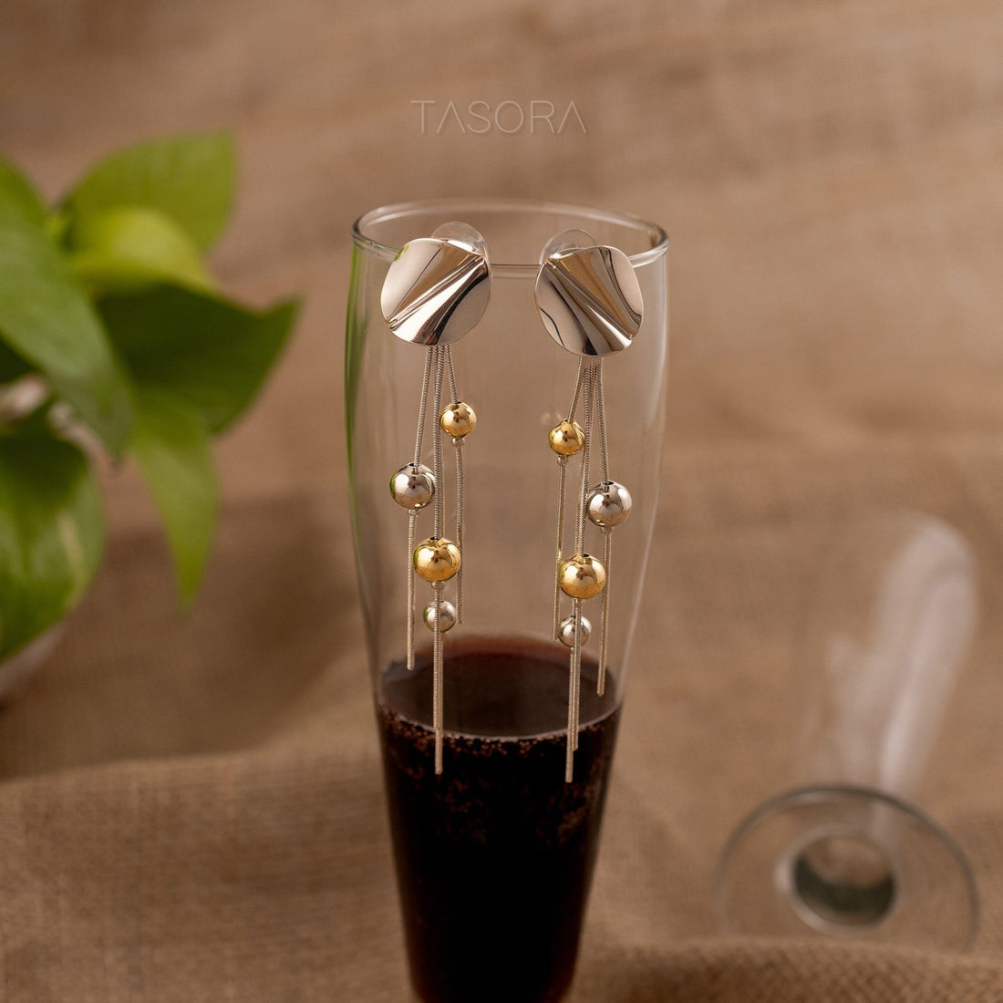 Glass with Disco Drizzle earrings by TASORA, placed on a textured surface with a plant in the background