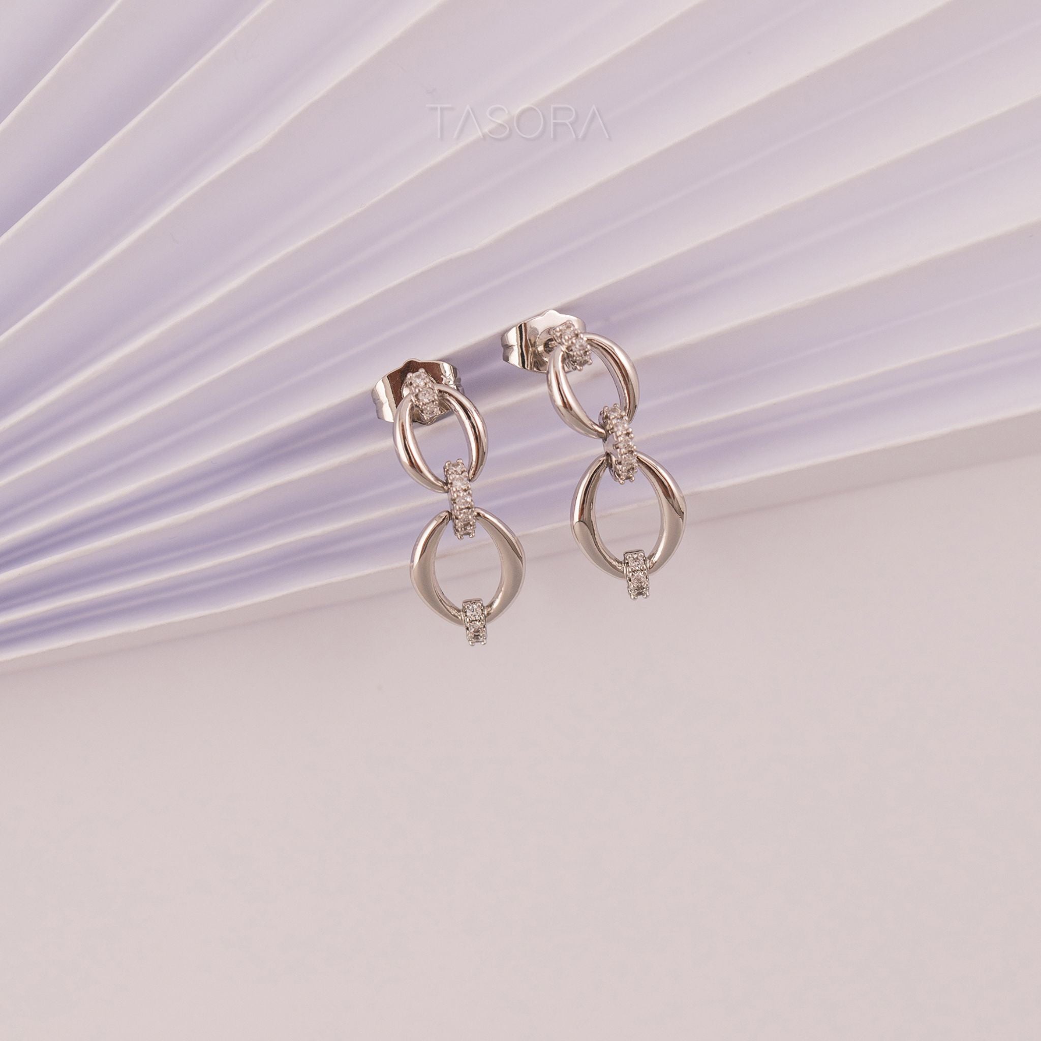Pair of Elior silver earrings on a light purple and white striped background with 'TASORA' branding.