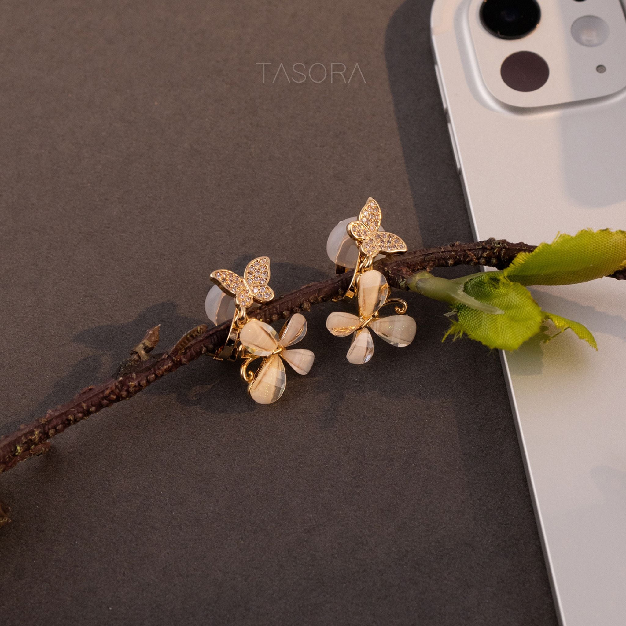 Golden Flutter Petal earrings on a branch with a phone in the background, branded 'TASORA'.