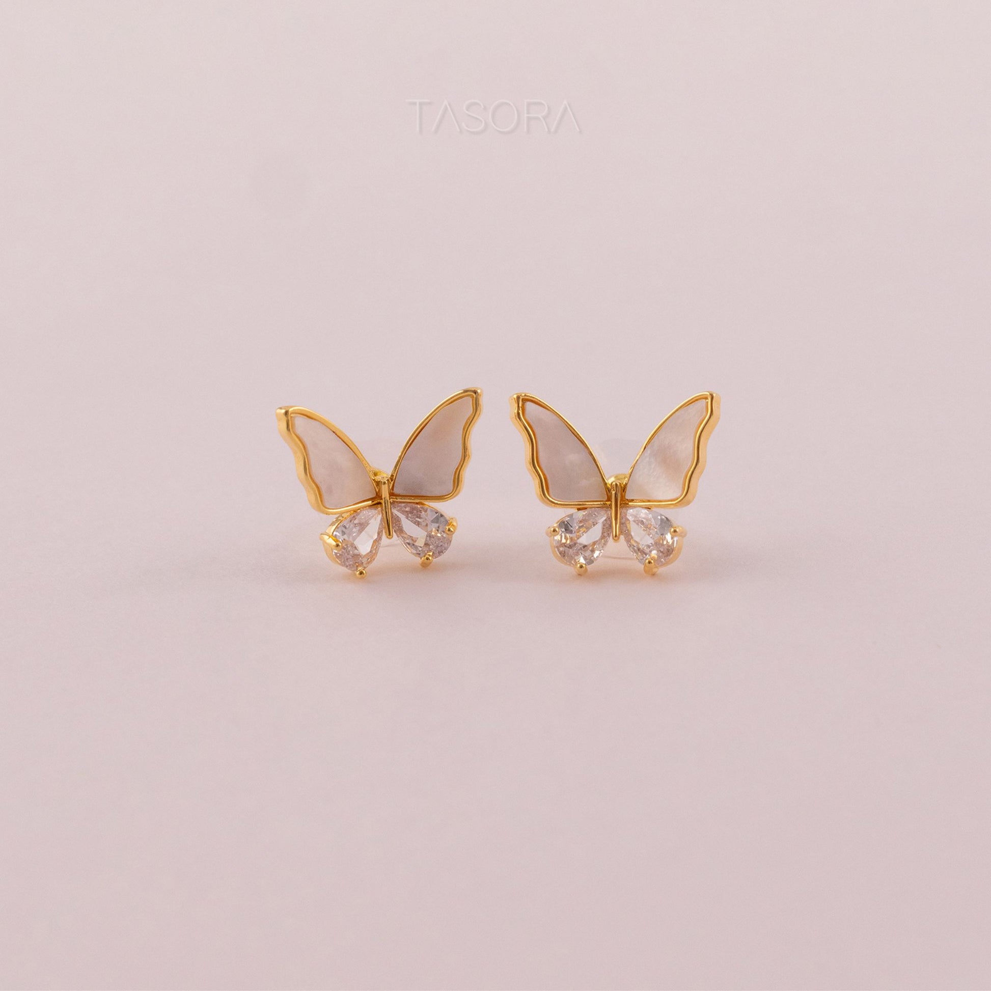Butterfly-shaped Flutter wings earrings of TASORA brand with gold frames and clear gemstones on a light pink background.