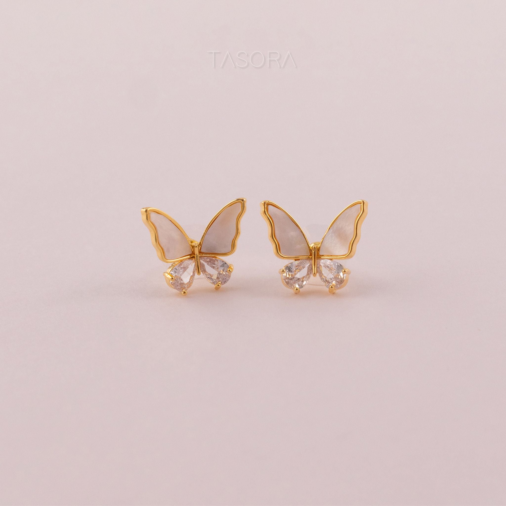 Butterfly-shaped Flutter wings earrings of TASORA brand with gold frames and clear gemstones on a light pink background.