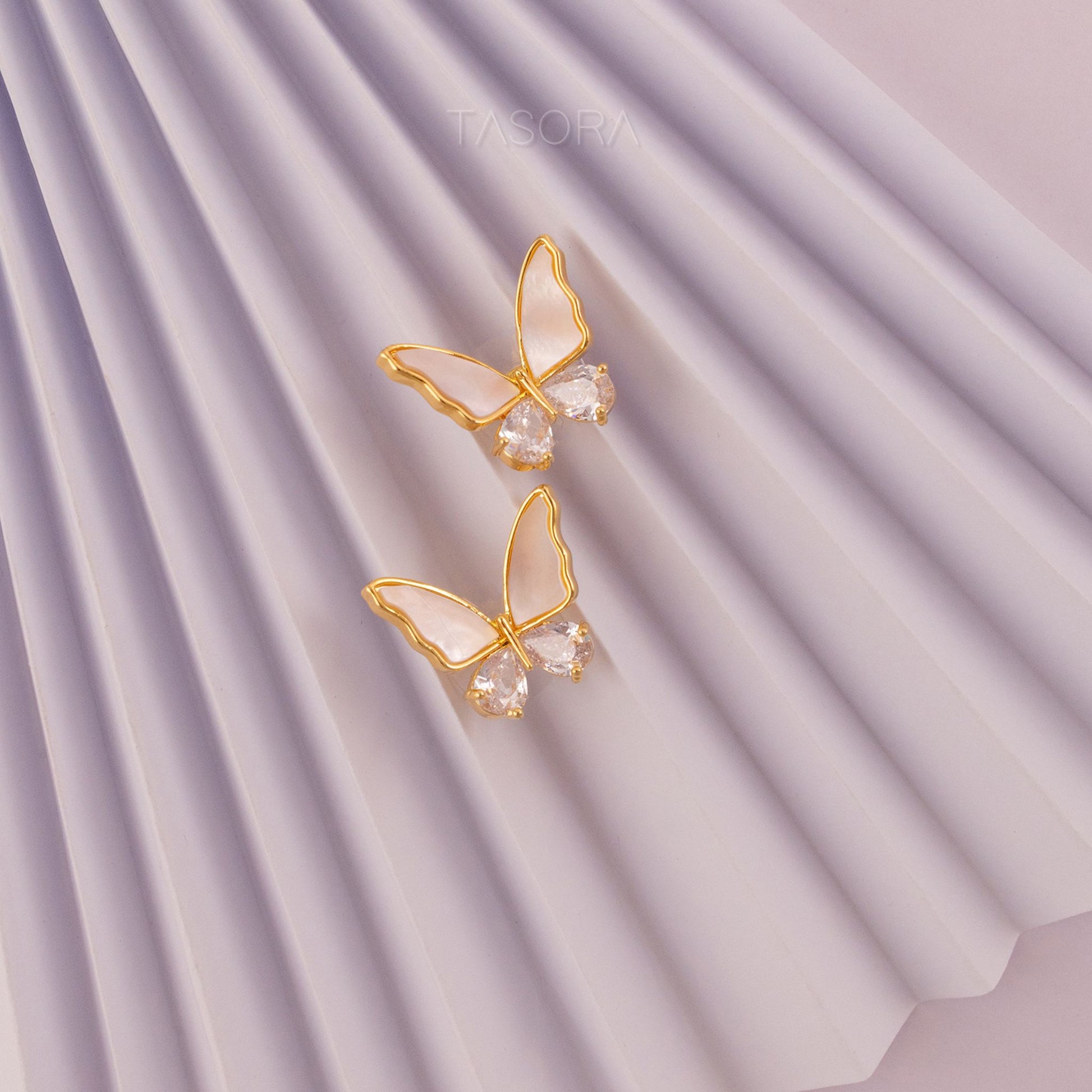 Butterfly-shaped Flutter wings earrings of TASORA brand with golden frames and clear gemstones on a Paper background.