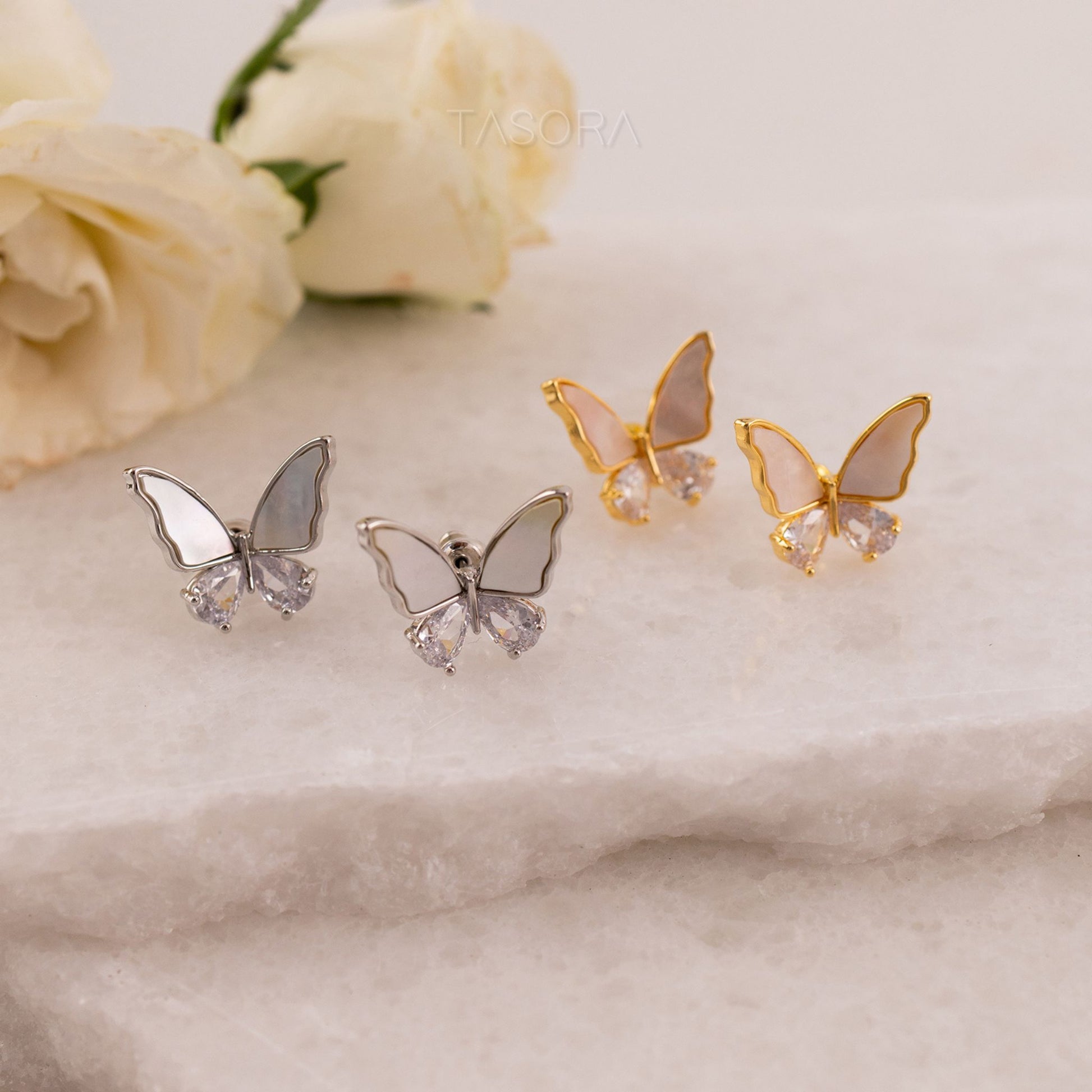 Butterfly-shaped Flutter wings earrings in gold and silver on a marble surface with white flowers in the background with TASORA branding