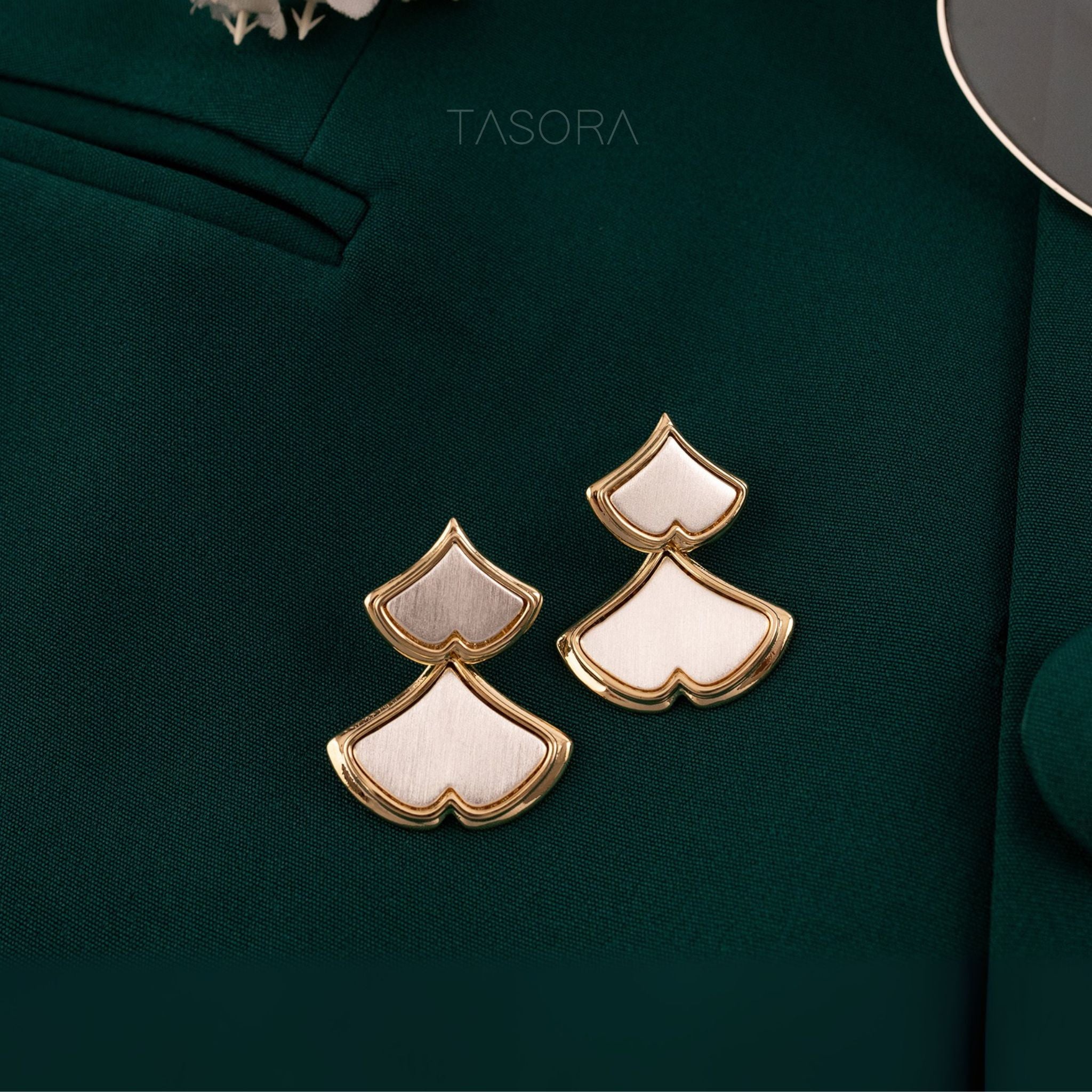 Pair of gold and white Golden Glint earrings on a dark green fabric background with 'TASORA' branding.