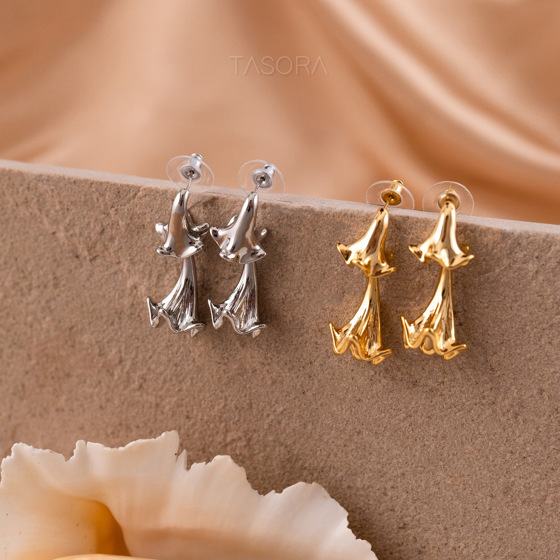 Pair of silver and golden Hibelle Drop earrings on a beige surface with 'TASORA' branding.