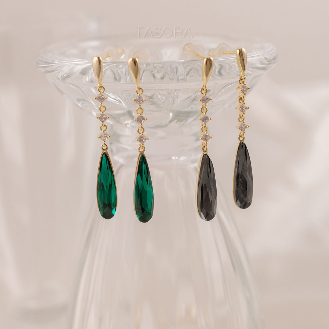 Pair of Noor Drop golden earrings with green and black teardrop-shaped stones on a clear stand with TASORA branding
