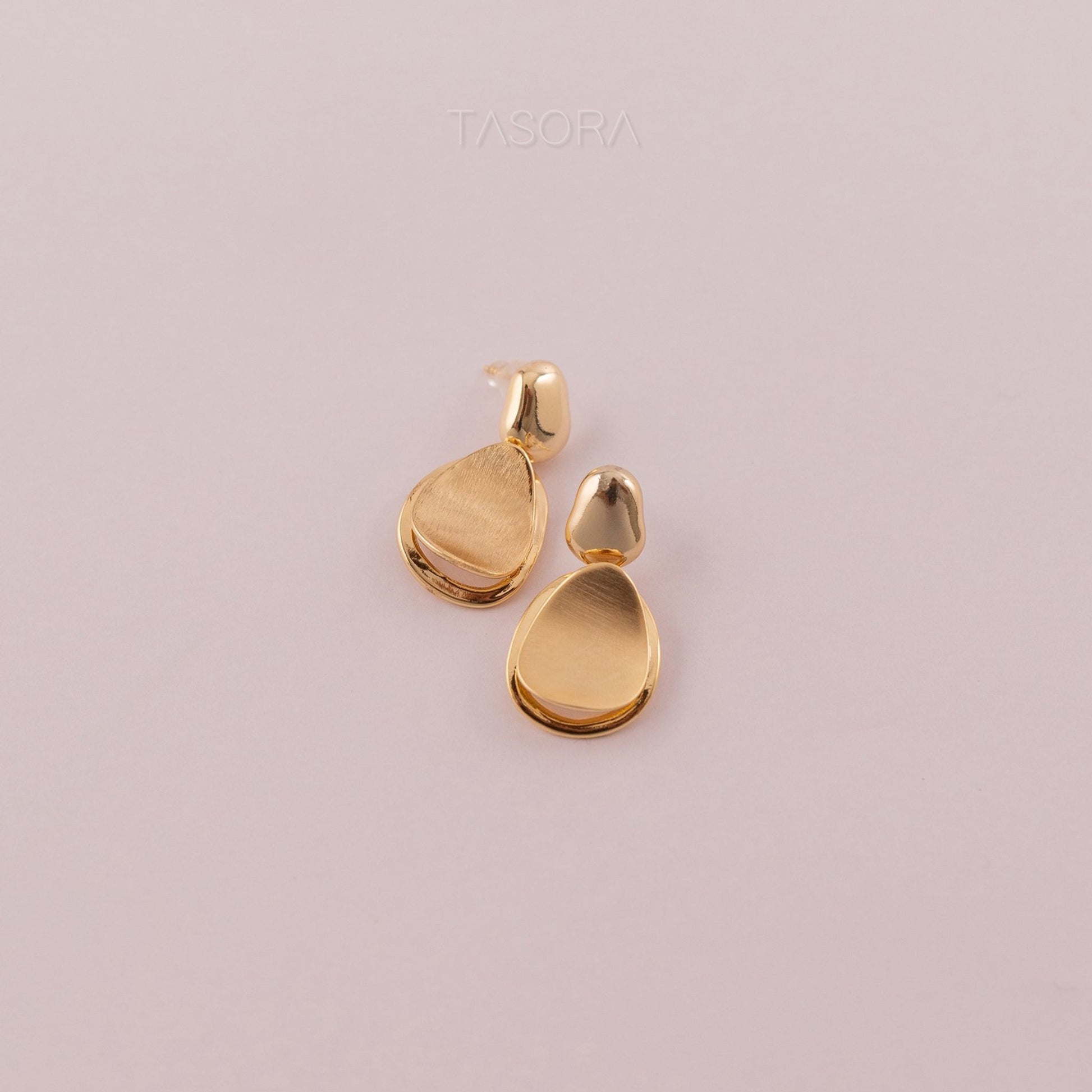 Gold teardrop-shaped earrings on a light pink background with 'TASORA' branding.