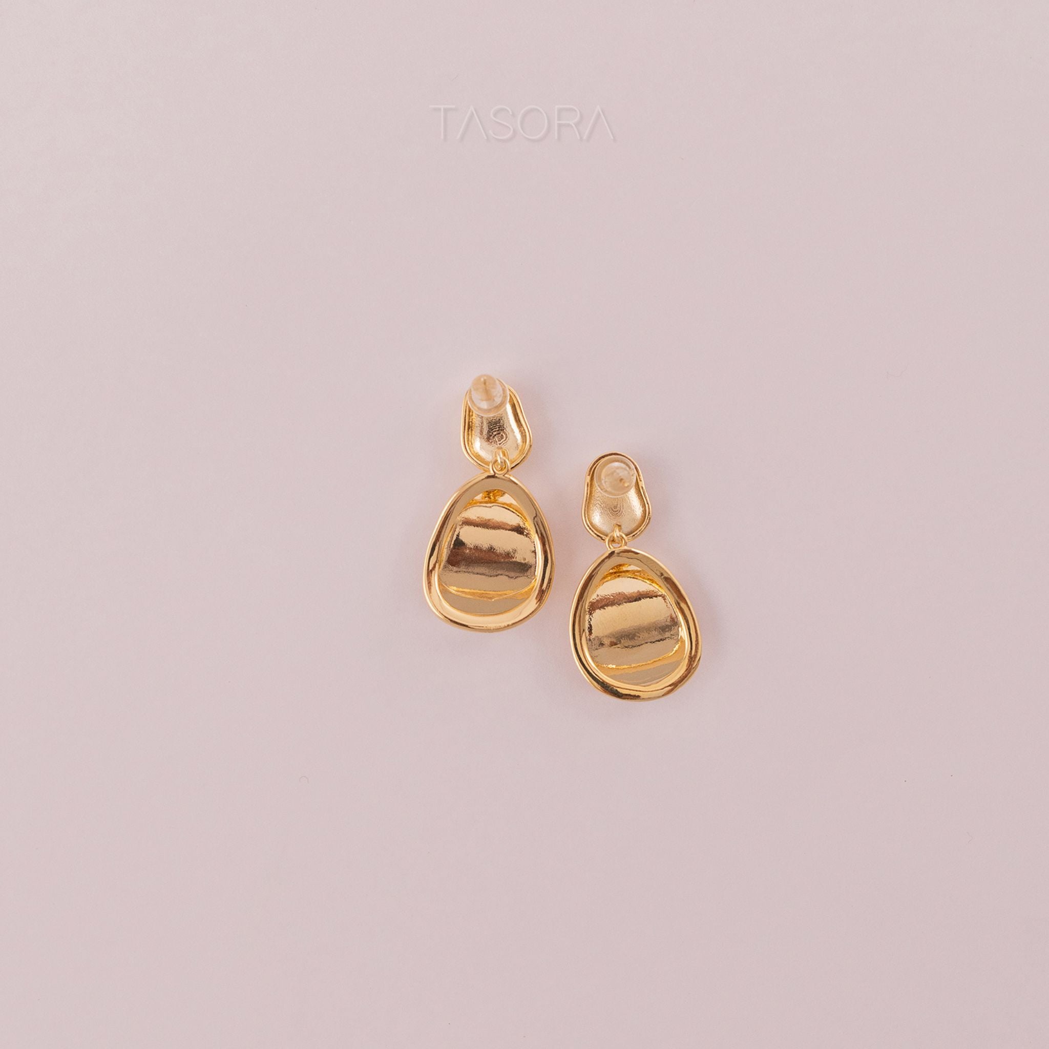 Back side of golden teardrop earrings on a light background with 'TASORA' branding.