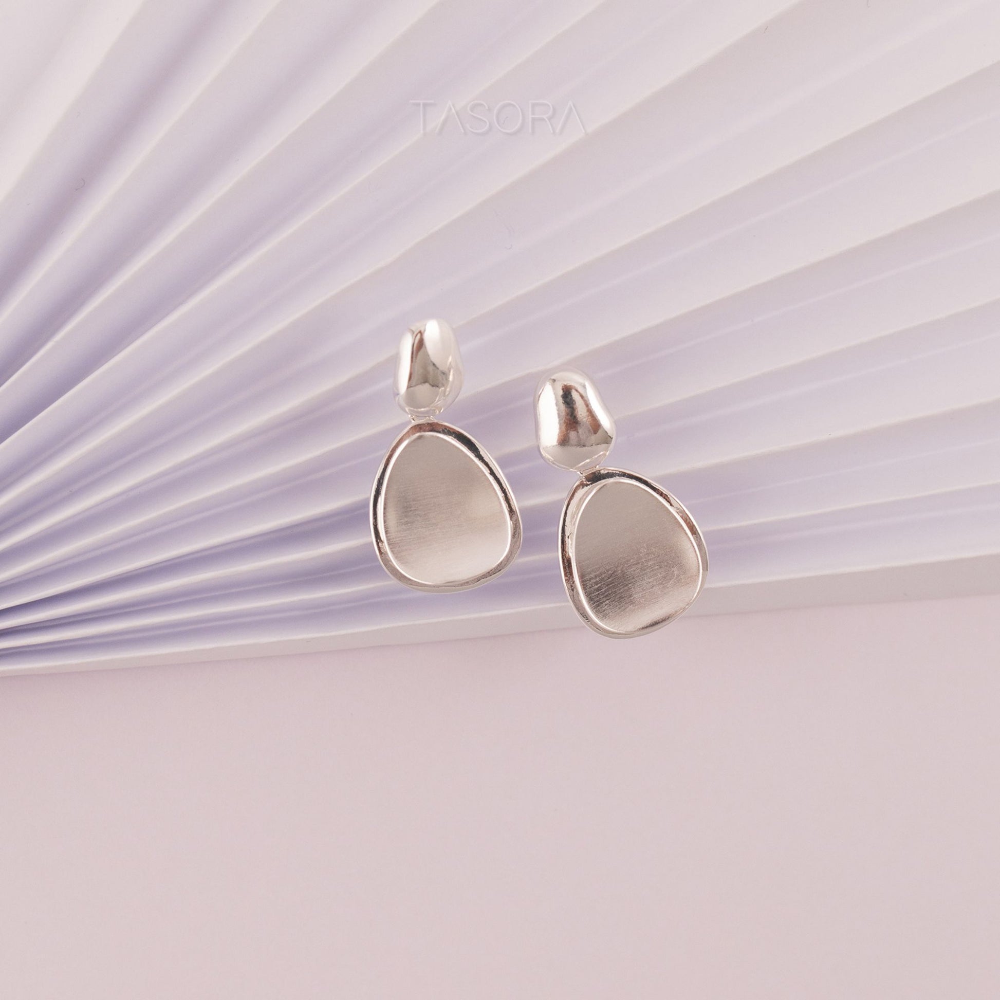 Pebble Huggie silver teardrop earrings on a light purple background with 'TASORA' branding.
