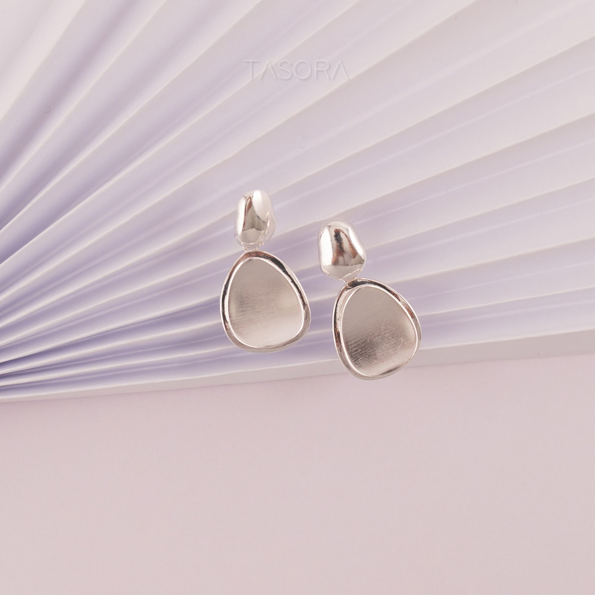Pebble Huggie silver teardrop earrings on a light purple background with 'TASORA' branding.
