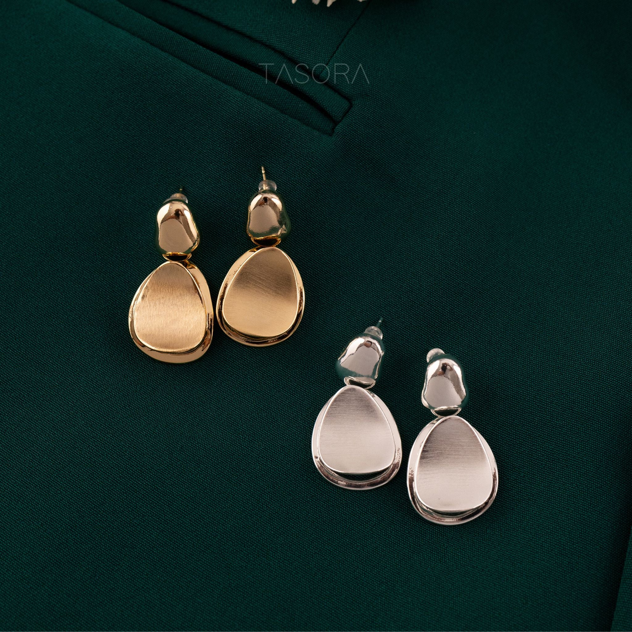 Gold and silver Pebble Huggie earrings on a dark green fabric background with 'TASORA' branding.