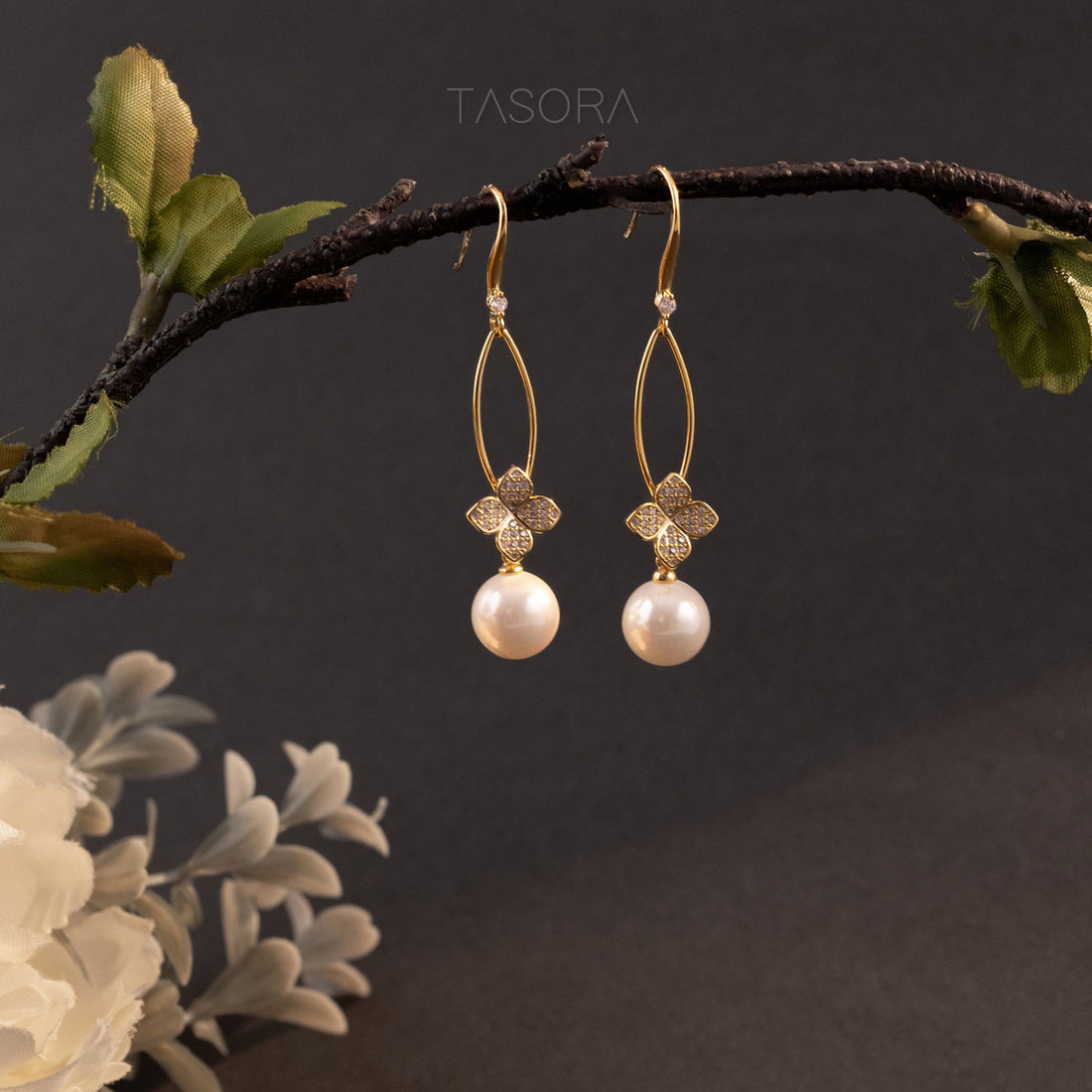 Golden Petal Fleur earrings with pearl drops on a branch against a dark background with TASORA branding