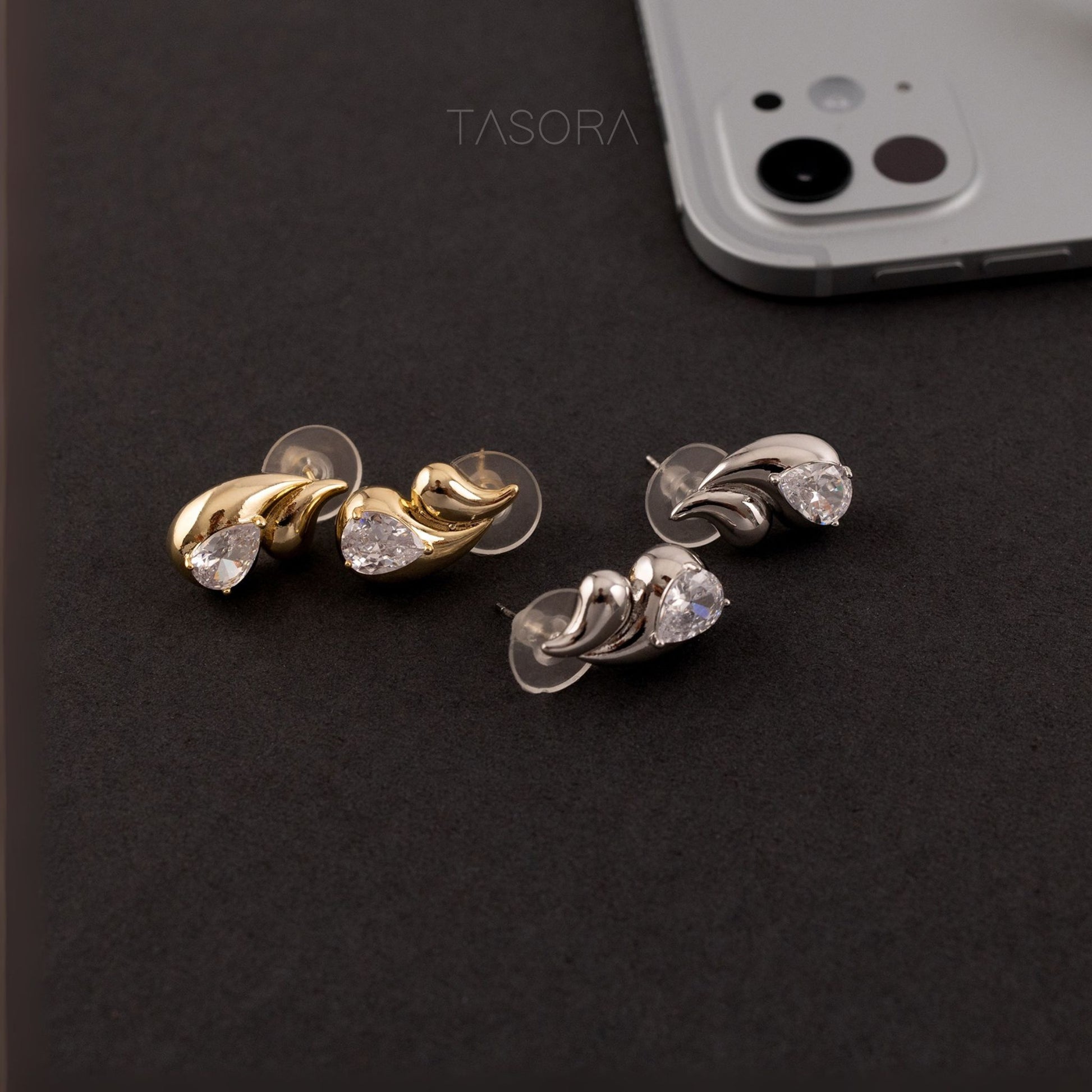 Pair of gold and silver Rain Drop earrings on a dark surface with a phone and TASORA branding in the background.