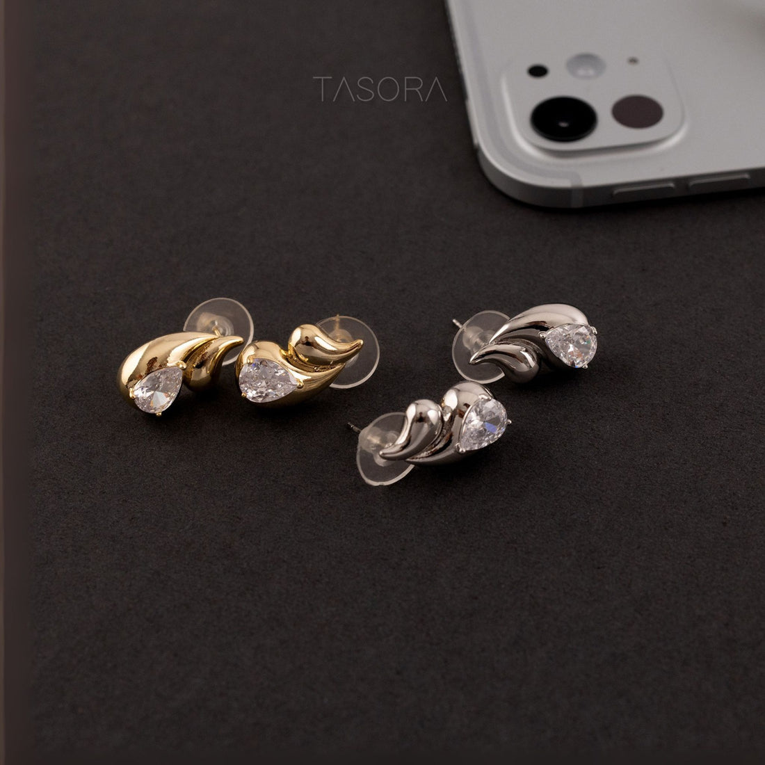 Pair of Rain Drop golden and silver earrings on a dark surface with a phone and TASORA branding in the background.