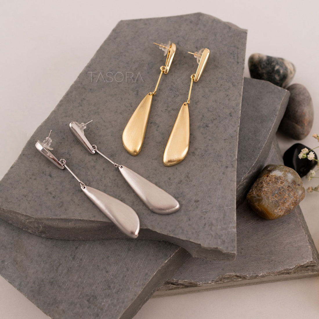 Gold and silver Sleeky Drop earrings on a stone surface with 'TASORA' branding.