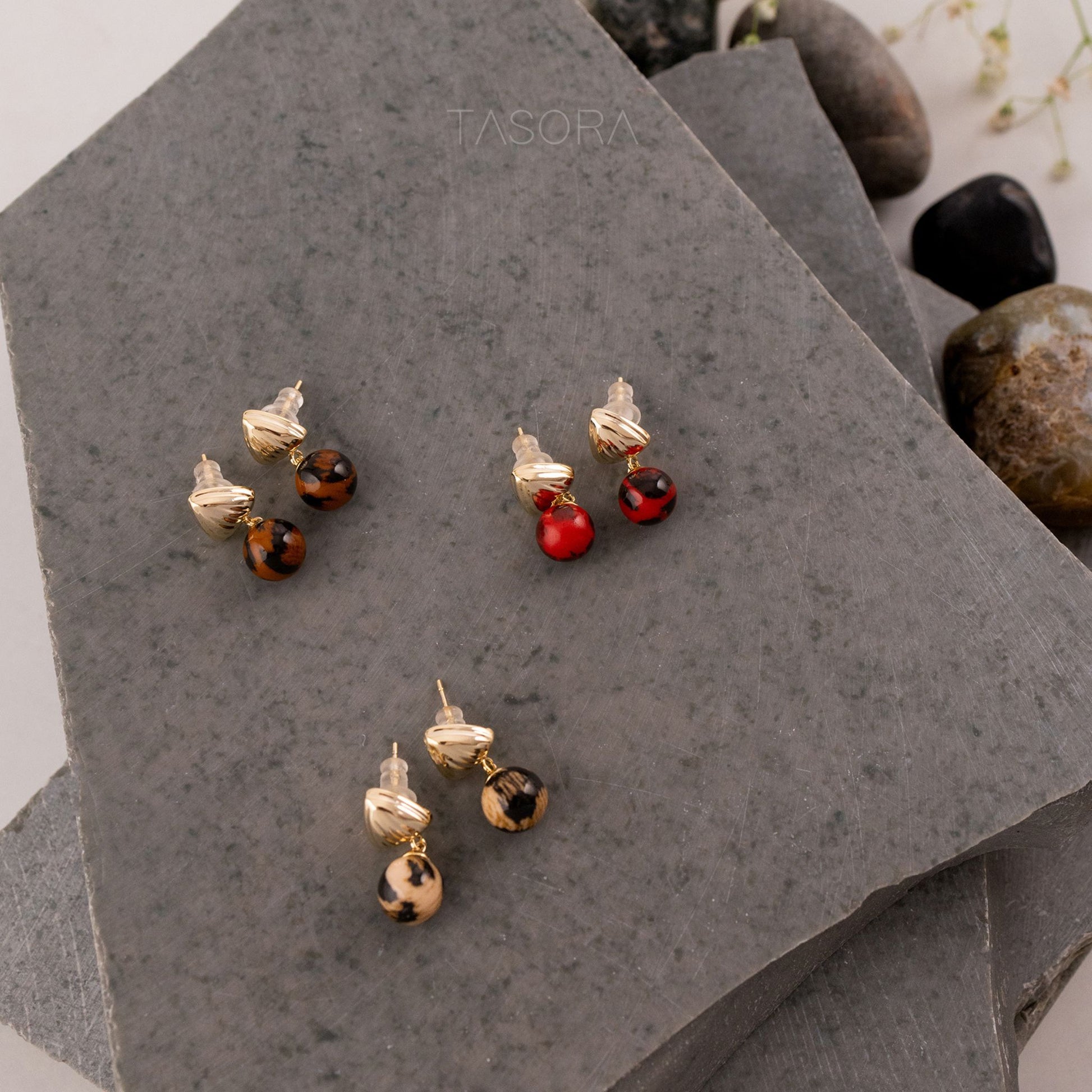 Pairs of Sona Halo earrings with red, brown and beige colour on a gray stone surface with TASORA branding