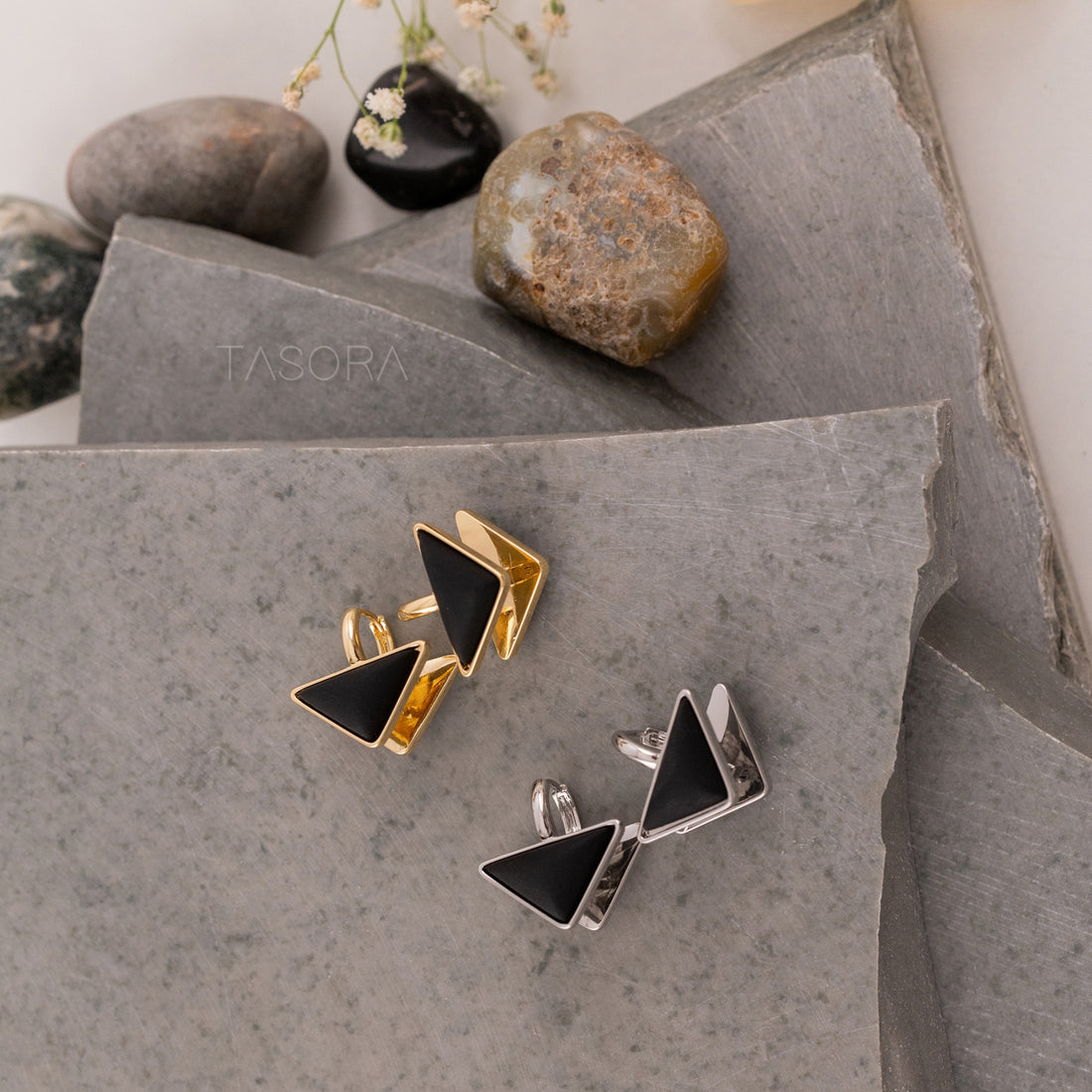 Pair of gold and silver triangle-shaped Trique Stud earrings on a stone surface with 'TASORA' branding.