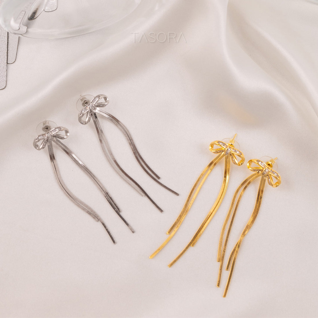 Silver and golden Twinkle Tie earrings by TASORA with bow details on a white fabric background