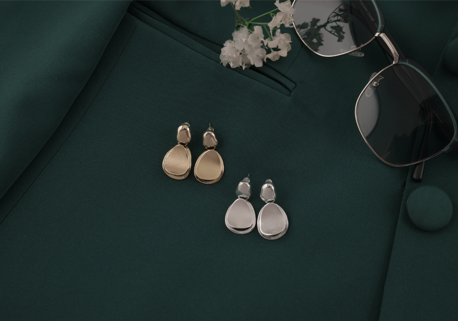 Pair of gold and silver earrings on a dark green surface with sunglasses and flowers.